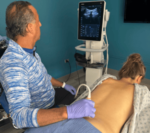 Dr. Cammisa wearing gloves and a blue patterned shirt performs an ultrasound scan on a patient's back, using a medical device while viewing the scan on a monitor.