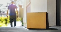 A close-up of a cardboard package left on a doorstep, with a delivery person walking away in the background. The image emphasizes the delivery of online purchases, possibly representing e-commerce platforms like Amazon or eBay.