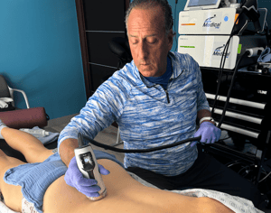 Dr. Cammisa, dressed in a blue and white striped shirt and purple gloves, is using a shockwave device on the lower back of a patient lying on a treatment table. The patient is partially covered with a hospital gown and is positioned to receive treatment in a clinical setting.