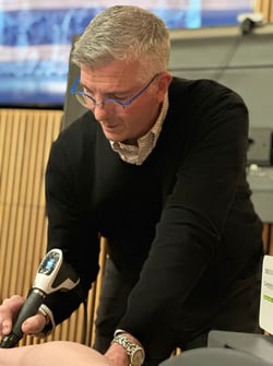 Dr. Conenello performing a treatment on a patient using a shockwave device. He is focused on the procedure, wearing a black sweater and glasses, with the device aimed at the patient's body.