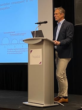 A presentation by Dr. Gerdesmeyer at a podium, explaining scientific concepts with a projected slide in the background.