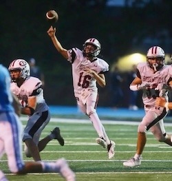 A high school quarterback in a white and maroon Ridgewood uniform, number 16, is captured mid-throw during a nighttime football game. He is surrounded by teammates in similar uniforms, with one to his left and another to his right. The background is dark, highlighting the players in action.