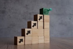 A wooden staircase made of small blocks, each with black arrows pointing upward. At the top of the staircase is a green block with a target symbol and a dart hitting the center, symbolizing progress and making decisions. 