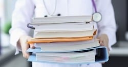 The image shows a medical professional wearing a white coat and stethoscope, holding a large stack of books and folders. The focus is on the hands and the materials, emphasizing research, documentation, or study, which may be related to medical evidence.