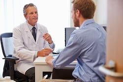 A middle-aged male doctor wearing a white lab coat is sitting at a desk, attentively talking to a male patient across from him. The doctor has a pen in his hand, ready to take notes. 