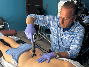 Dr. Cammisa uses a handheld device to apply treatment to a patient's lower back, while the patient lies on a treatment table with a towel covering part of their body.